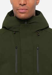 Giacca verde oliva con colletto alto, dotata di due tasche verticali con zip e dettagli con cordino. Texture liscia con un design moderno.
