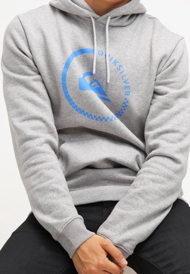 Personne portant un sweat à capuche gris clair avec un logo Quiksilver bleu, assise les mains jointes sur un pantalon noir sur un fond blanc.