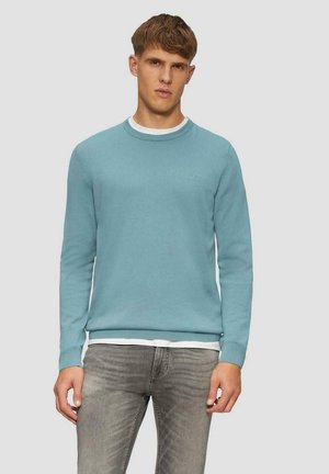 Lichtblauwe trui van zachte stof, met een ronde hals en geribde boorden. Draagt over een wit T-shirt en gecombineerd met grijze jeans.