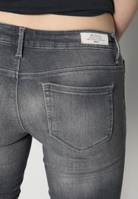 Vaqueros de denim gris con un acabado desgastado, que cuentan con bolsillos traseros y una etiqueta de marca en la cinturilla. Vista en primer plano desde la parte trasera.