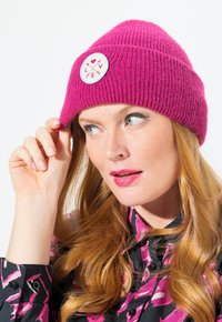 Gorro de punto fucsia con textura acanalada y un parche redondo blanco que presenta un corazón rosa y flechas. Resaltado con un diseño juguetón.