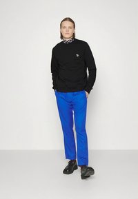 Sweat-shirt noir avec un petit logo, associé à un pantalon bleu vif, porté avec des chaussures noires. Design minimaliste, texture lisse et coupe ajustée.