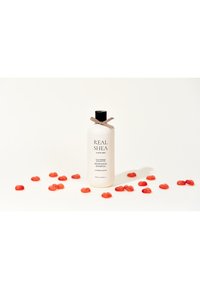 Bottiglia di shampoo bianca con tappo nero e nastro marrone, etichettata "REAL SHEA" e circondata da caramelle a forma di cuore rosso su uno sfondo bianco.