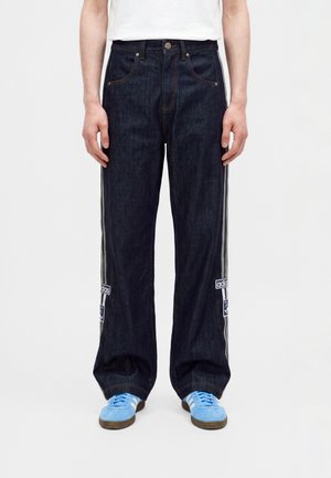 Person iført mørkeblå vidde jeans med sidelister og Adidas-patches, kombineret med lyseblå sneakers og en hvid top.