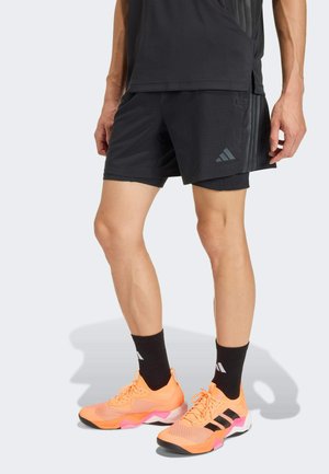 Mann trägt schwarze Sportshorts, schwarze Socken und orangefarbene Laufschuhe, steht vor hellem Hintergrund.