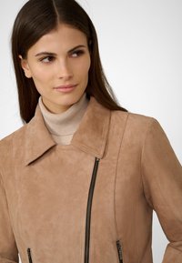 Beige mockaskinnjacka med spetsig krage, diagonala blixtlås och figurnära design. Buren över en ljus polotröja. Slät textur, minimal detaljering.