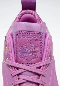 Gros plan de la languette rose d'une basket Reebok Classic avec logo embossé, coutures et lacets attachés sur fond blanc.
