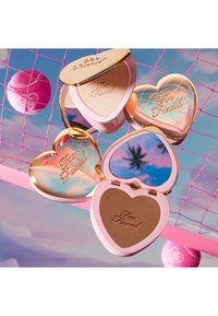 Hartvormige compacte spiegel, roze en goud, met de gegraveerde tekst "Too Faced." Bevat een reflecterende spiegel en bronzer. Soepele textuur, glanzende afwerking.