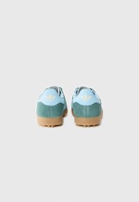 Baskets en daim vert avec des accents bleu clair et une semelle en caoutchouc beige. Caractérisées par le logo trèfle d'Adidas au niveau du talon.
