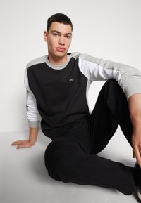 Felpa bloccata nei colori nero e grigio con maniche lunghe bianche, scollatura rotonda e piccolo logo verde. Indossata con pantaloni neri.