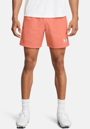 Atleta masculino con pantalones cortos coral de Under Armour, calcetines deportivos blancos y zapatos con tacos blancos, de pie contra un fondo liso.