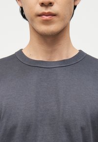 Κοντινό πλάνο του κάτω μέρους του προσώπου και του λαιμού ενός ατόμου που φοράει μια απλή σκουρόγρια μπλούζα με στρογγυλή λαιμόκοψη, σε ουδέτερο φόντο.