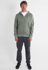Sweat à capuche vert zippé avec une doublure beige, poches avant et poignets côtelés, associé à un pantalon gris foncé et des chaussures noires.