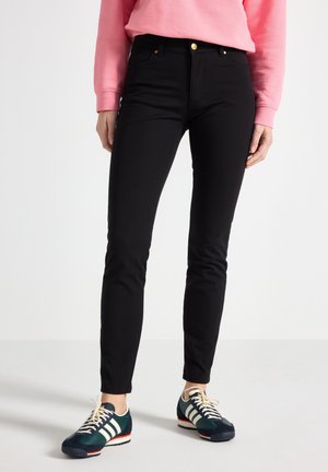 Person iført sorte slim-fit bukser, mørkegrønne og hvide sneakers og en lyserød langærmet top, stående på en ensfarvet lys gulv.