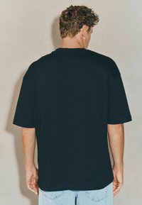 Black short-sleeve t-shirt with a loose fit and round neckline, made from cotton, shown from the back with visible seams. Light blue jeans are worn.