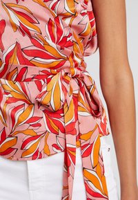 Rosa stofbluse med røde og orange bladmønstre, med en knyttet talje detalje og glat tekstur. Accenter inkluderer hvide kanter.