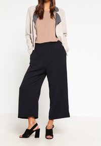 Lys grå cardigan med en marineblå krave, tan top, sorte brede bukser og sorte blokhæle sandaler. Glatte teksturer og løs pasform.