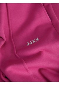 Un tejido fucsia con una textura suave presenta el texto bordado "JJXX" en blanco contrastante en la esquina inferior derecha.