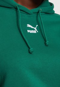 Sweat à capuche vert en coton, avec une poche kangourou, des cordons de serrage réglables et un logo Puma blanc sur la poitrine. Texture douce.