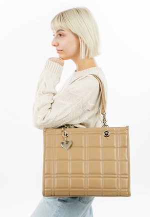Sac tote beige avec texture matelassée, quincaillerie métallique et charme en forme de cœur. Il dispose de poignées doubles et d'une forme rectangulaire structurée.