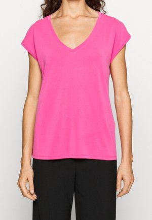 Kvinde iført en lys pink kortærmet V-hals top, kombineret med sorte bukser, vist fra halsen til hofterne mod en ensfarvet baggrund.