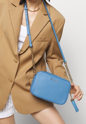 Vrouw met een beige blazer en witte kanten korte broek houdt een blauwe Michael Kors crossbodytas met gouden ketting en kwastdetail vast.