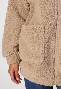 Beige fleecová bunda s texturovaným, chlupatým povrchem, která má přepychové kapsy a zipové zapínání. Rukávy a lem jsou také měkké.