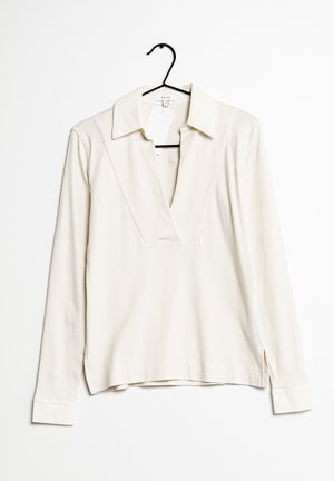 Krémové polo tričko s dlouhým rukávem a hlubokým výstřihem do V zavěšené na černém ramínku na bílém pozadí.