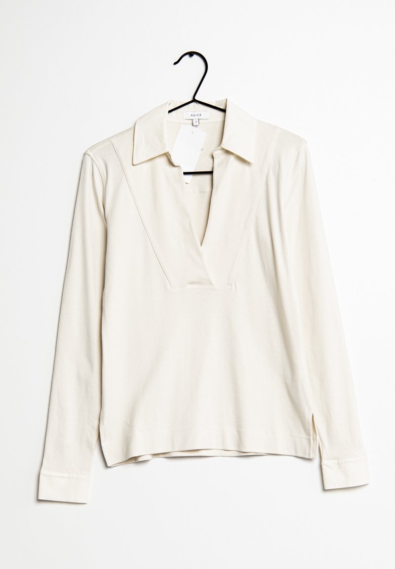 Polo à manches longues crème avec un col en V profond, suspendu sur un cintre noir contre un fond blanc.