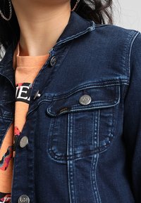 Close-up of person wearing dark blue denim jacket over orange graphic T-shirt, showing jacket buttons and collar details.
