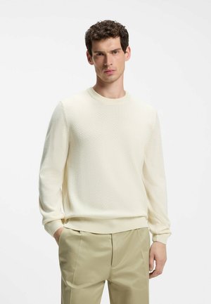 Junger Mann mit kurzen lockigen Haaren, der einen cremefarbenen, strukturierten Pullover und beige Hosen trägt, steht mit einer Hand in der Tasche vor einem schlichten Hintergrund.