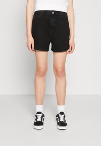 Shorts de mezclilla negra con dobladillo enrollado, diseño de cinco bolsillos y botón frontal. Llevados con zapatillas negras y calcetines blancos.