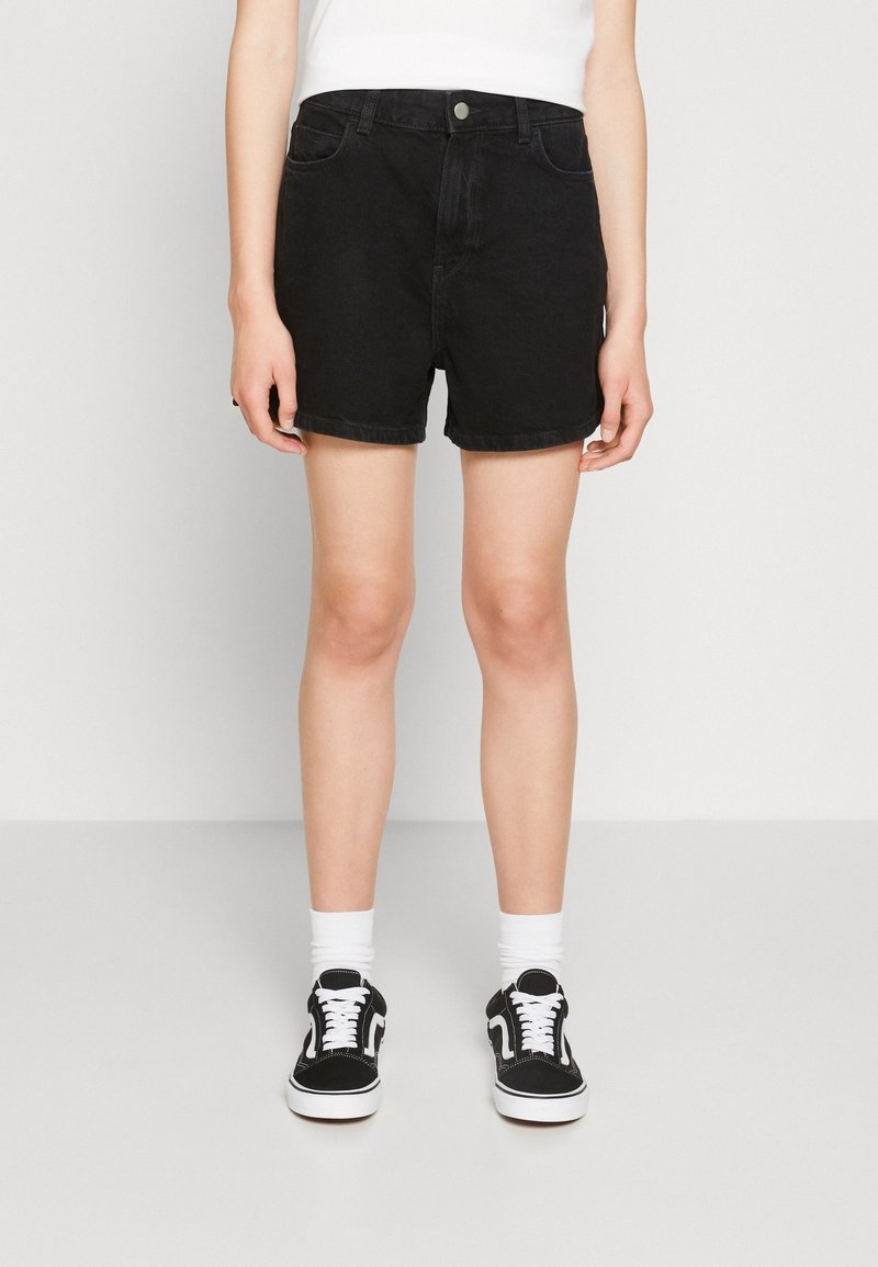 Shorts de mezclilla negra con dobladillo enrollado, diseño de cinco bolsillos y botón frontal. Llevados con zapatillas negras y calcetines blancos.