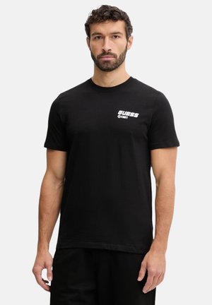 Uomo con capelli ricci e barba che indossa una T-shirt nera Guess 1981, in piedi contro uno sfondo chiaro e uniforme.