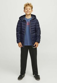 Námornícká puffer bunda s kapucňou, rebrovanými manžetami, zapínaním na zips, nosená cez modré tričko a čierne tepláky, v kombinácii s čiernymi teniskami.