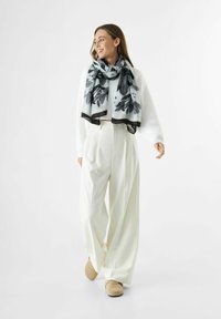 Foulard à motifs floraux noir et blanc sur un pull blanc. Le modèle porte un pantalon large blanc et des chaussures beiges, sur un fond clair.