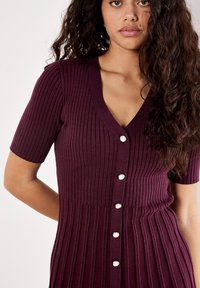 Donna con lunghi capelli ricci che indossa un cardigan corto a coste di colore prugna con bottoni di perle, in posa davanti a uno sfondo semplice.
