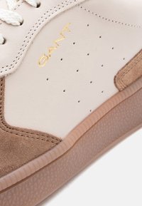 Λευκό και μπεζ αθλητικό παπούτσι πλαϊνή όψη με χρυσό λογότυπο "GANT", διάτρητο επάνω μέρος, λευκά κορδόνια και υφασμένη ελαστική σόλα ανοιχτού καφέ χρώματος.