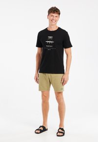 Zwart T-shirt met korte mouwen en witte graphic print, gecombineerd met lichtgroene shorts en zwarte sandalen. Effen achtergrond.