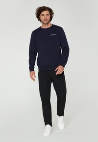 Sudadera de algodón azul marino con cuello redondo y mangas largas, que presenta una impresión de logo blanca en el pecho; combinada con pantalones negros ajustados y zapatillas blancas.
