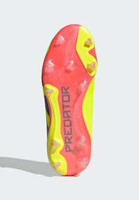 La semelle de la chaussure de football présente un design vibrant en rouge, jaune et noir avec une surface texturée et des crampons placés de manière stratégique pour une traction optimale.
