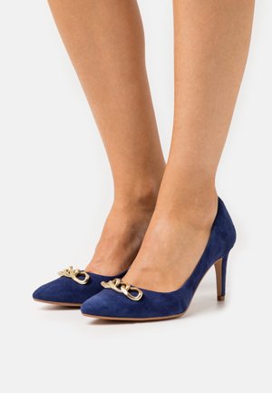 Pompes à talons hauts en daim bleu marine avec des pointes et des accents décoratifs en chaîne dorée à l'avant. Texture lisse et talons fins.