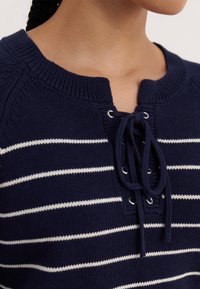 Mörkblårandig tröja med vita horisontella ränder, med en snörning i halslinningen och ribbad stickad struktur. Kort, rundad krage.