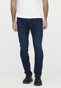 Jean en denim bleu foncé ajusté avec un léger délavage, cinq poches et des détails de surpiqure contrastants, associés à des baskets noires.