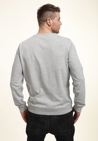Felpa grigia con maniche lunghe, collo a giro e polsini a costine. Tessuto liscio e vestibilità rilassata, indossata sopra jeans scuri, vista da dietro.