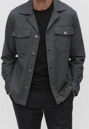 Mann trägt eine dunkelgraue Jacke mit Brusttaschen über einem schwarzen Hemd, Hände in den Taschen, steht vor einem schlichten Hintergrund.