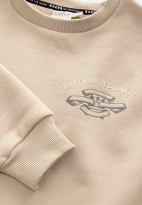 Beige sweatshirt med ribbade ärmslut och halsringning. Har ett grafiskt tryck i grått med texten "BAKER BY TED BAKER" och en logotypdesign.