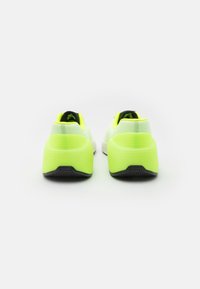 Un paio di sneaker neon gialle e bianche viste da dietro su uno sfondo bianco, evidenziando le suole spesse e le suole nere.