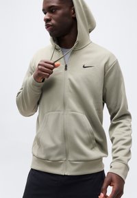 Άνδρας που ρυθμίζει τη κουκούλα μιας ανοιχτόχρωμης πράσινης ζακέτας Nike με φερμουάρ, φοράει μαύρο παντελόνι, μπροστά σε απλό ανοιχτόχρωμο φόντο.