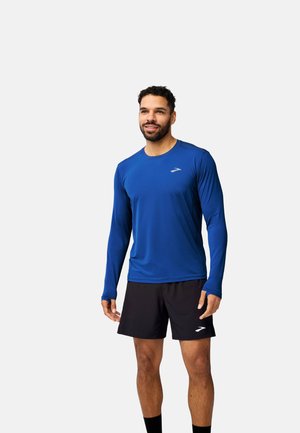 Langärmliges blaues Sportshirt aus leichtem Material, kombiniert mit schwarzen Shorts. Verfügt über ein kleines Logo auf der Brust.